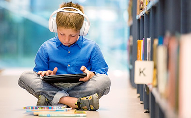 Child sitting on a library floor listening to headphones while using an iPad